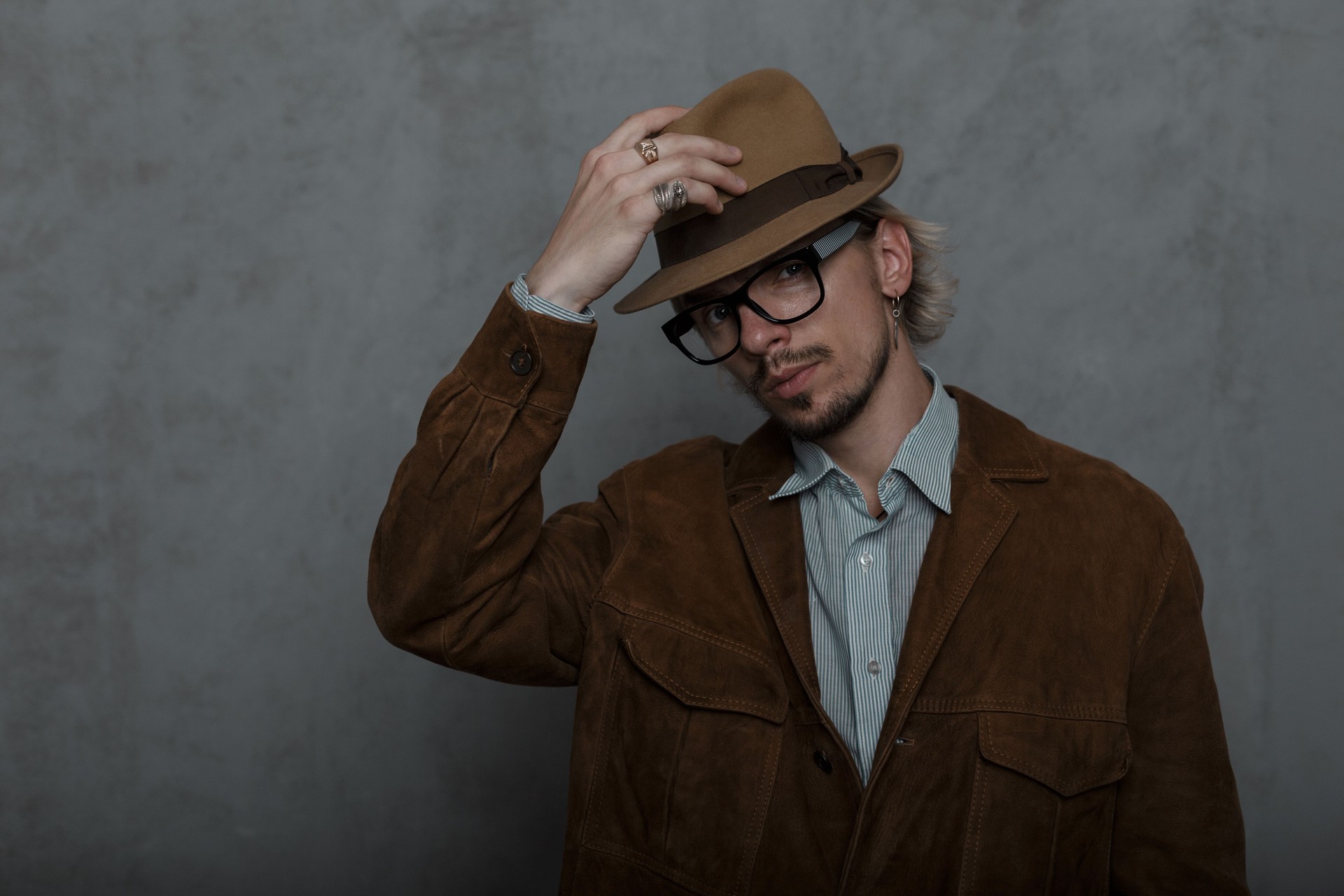 Old-fashioned young man hipster with a beard straightens a stylish hat in a room near a gray wall. Sexy guy in vintage glasses in a brown fashionable jacket in a classic shirt posing indoors. Retro.