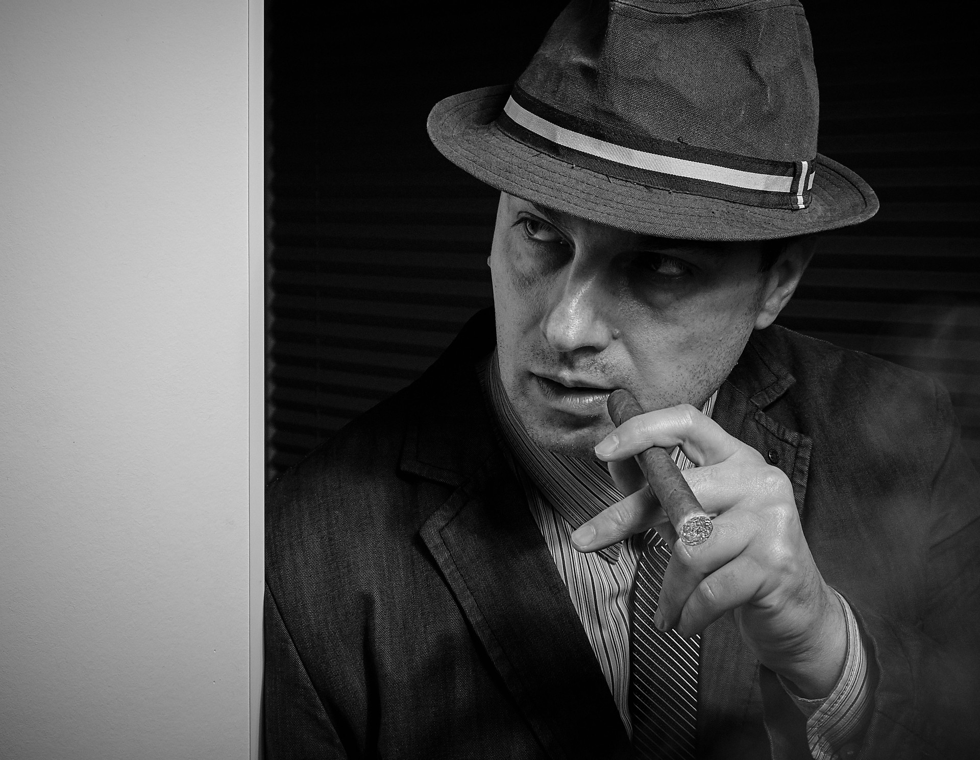 A mysterious man in a fedora and suit stands in a dark room, holding a cigar